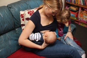 Mother feeding baby in underarm/rugby hold while toddler watches from other side