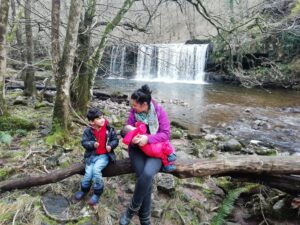 Mum sat and nursing a baby with an older child on a tree branch in the woods, with a waterfall behind